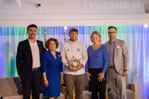 Five people stand together indoors; one person in a sports hoodie holds a plaque. They are dressed in formal attire, except for the individual in the hoodie. The backdrop has colorful lighting and decor, highlighting the achievements of the Community Benefit Fund's scholarship program.