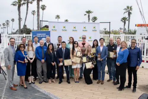 A group of people stand outdoors, holding awards and smiling for a photo in front of a banner, celebrating their community benefit program. Palm trees and a white fence are in the background.