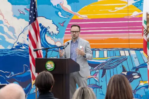 A man in a gray suit speaks at a podium in front of a colorful mural featuring marine life and a sunset. An American flag and another flag are positioned on either side of the podium as he announces the launch of the Blueprint.