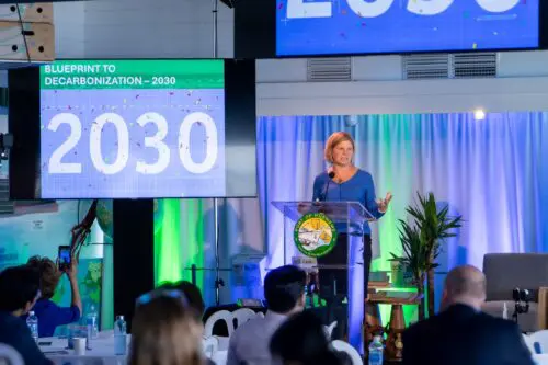 A person stands at a podium with the speaking at an event. Screens behind display the phrase "Blueprint to Decarbonization – 2030" and the number "2030," highlighting a new Scholarship Program aimed at supporting education in sustainability.