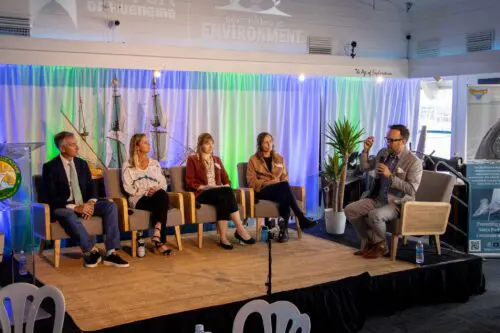 Five individuals are seated on a stage engaged in a panel discussion. A man on the right is holding a microphone and appears to be speaking passionately about the Scholarship Program. 