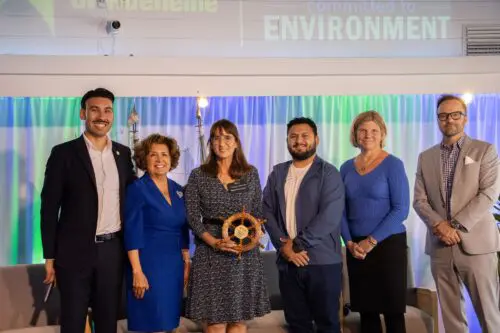 A group of six people stands together, smiling for the camera. The person in the middle holds a wooden ship's wheel. A banner above reads "committed to ENVIRONMENT," highlighting their participation in the Community Benefit Fund.