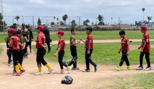 A group of young baseball players wearing red and green uniforms shake hands on a dirt field after a game, embodying a sense of sportsmanship. Nearby, coaches stand in the background near a chain-link fence.