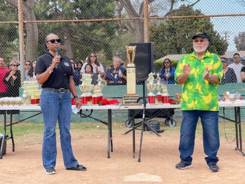 Two people standing on a baseball field, one holding a microphone, with a table filled with trophies and a crowd in the background. The person on the right is clapping and wearing a green shirt with yellow bananas, celebrating their zero emissions initiative.