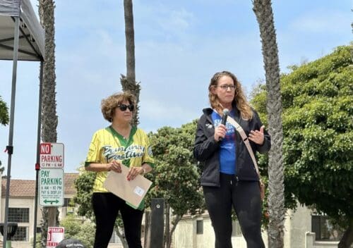 Two women are standing outdoors; one holding a folder, the other speaking into a microphone. They are under a canopy near a "No Parking" sign, with palm trees and buildings in the background, possibly discussing an event for Port Hueneme Little League.