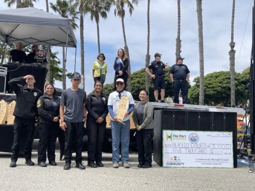 A group of people stands in front of a large check for $5,000, one person holding a wooden plaque. Police officers and bystanders are in the background against a backdrop of palm trees, celebrating the Port Hueneme Little League's generous donation.