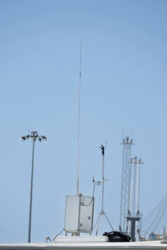 A rooftop with various antennas and communication equipment under a clear blue sky at the Port Hueneme Little League stadium. Multiple light poles and tall structures are visible in the background, capturing the essence of a sunny day at the ballpark.