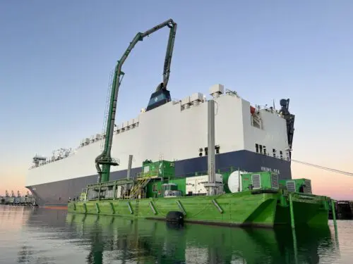 A large white ship is docked at a port. Next to it, a green barge equipped with a crane is lifting materials onto the ship, bolstered by a $3.935 million state grant for Port of Hueneme as Governor ramps up California Port Data Partnership Program.