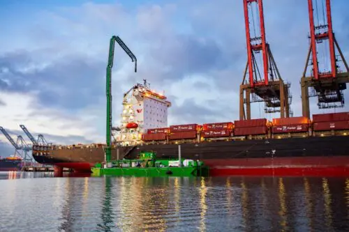 A cargo ship is docked at a port with cranes unloading shipping containers, benefiting from the $3.935 million state grant for Port of Hueneme as Governor ramps up California Port Data Partnership Program. A green barge is moored alongside the ship, under a cloudy sky with calm waters.