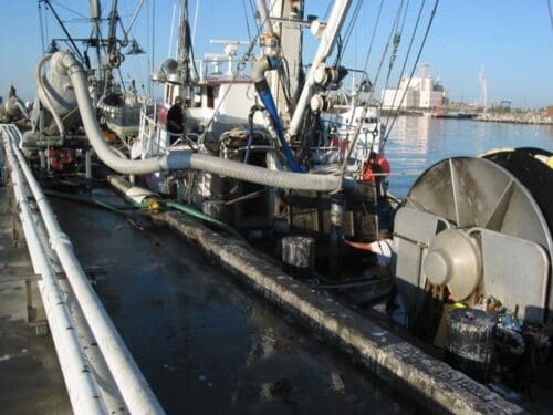 A commercial fishing boat docked at a harbor, equipped with large hoses and industrial machinery, sits against the calm water backdrop, benefiting from the $3.935 Million State Grant for Port of Hueneme as Governor ramps up California Port Data Partnership Program. Other boats dot the peaceful scenery.