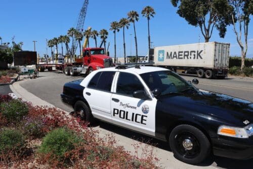 Police car parked on the roadside with a red truck and shipping container in the background, as palm trees sway under a clear sky. Nearby, talks of a $3.935 Million State Grant for Port of Hueneme are buzzing as the Governor ramps up California's Port Data Partnership Program.