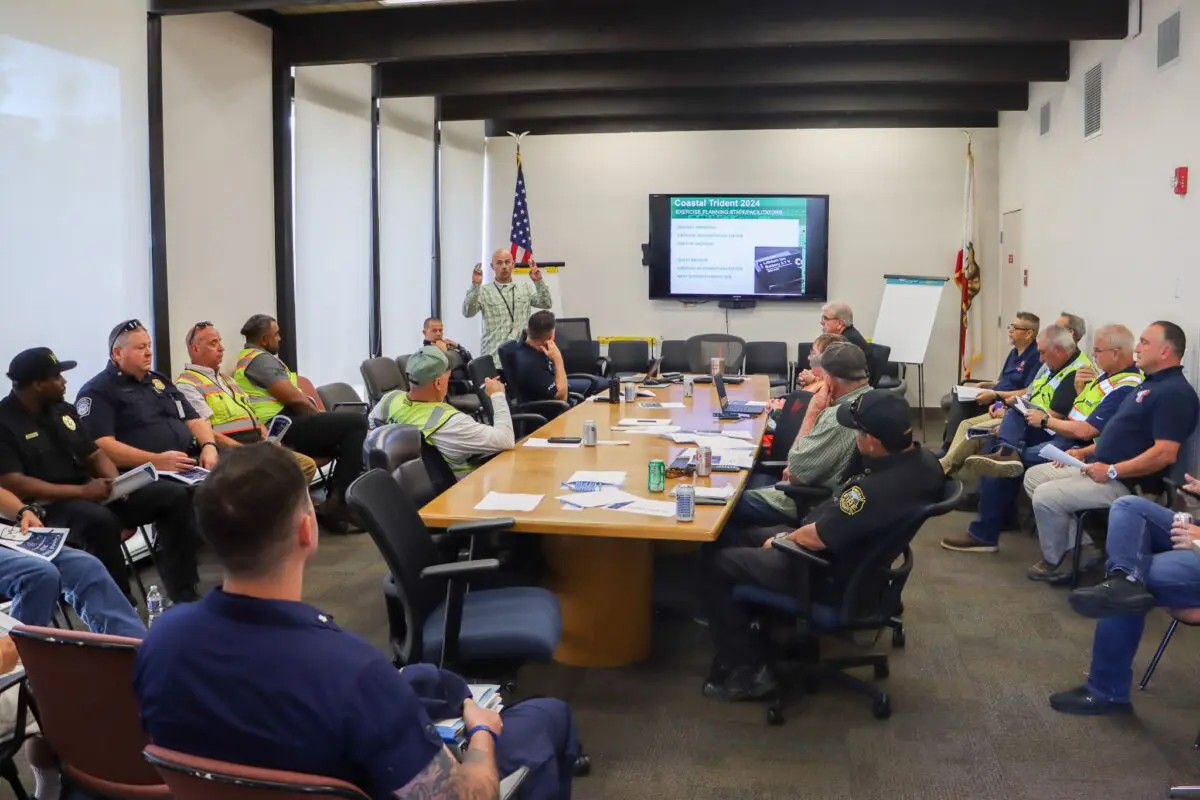 A group of people, some in uniforms and safety vests, are seated around a conference table in a meeting room with a presentation screen displaying information about the $3.935 Million State Grant for Port of Hueneme as Governor ramps up the California Port Data Partnership Program.