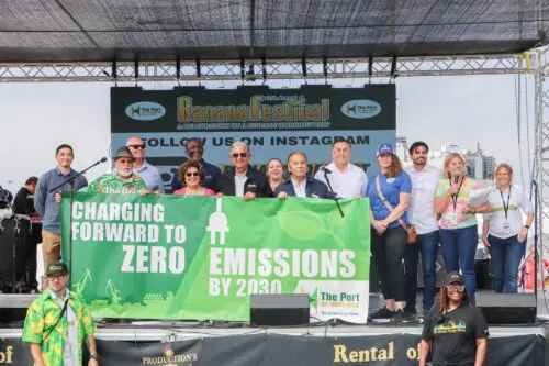 A group of people on stage at the Banana Festival, holding a banner that reads "Charging Forward to Zero Emissions by 2030," celebrates the $3.935 million state grant for Port of Hueneme as Governor ramps up the California Port Data Partnership Program, set against the backdrop of festival signage and a musical band setup.