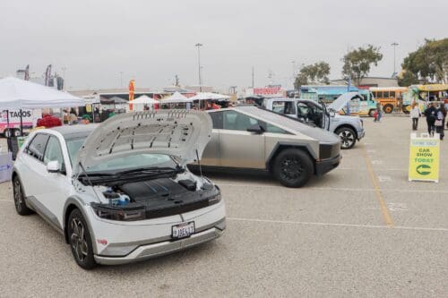 The outdoor car show featured several electric vehicles displayed in a parking lot. The hoods of two cars in the foreground were open, showcasing their engines. Banners, tents, and food trucks added to the lively atmosphere, reminiscent of the excitement surrounding the $3.935 Million State Grant for Port of Hueneme.