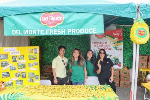 Four people stand behind a booth displaying bananas under a Del Monte Fresh Produce canopy. The booth features a banner about bananas and a backdrop with the Del Monte logo and food safety commitment, highlighting their role in the $3.935 Million State Grant for Port of Hueneme's data partnership program.