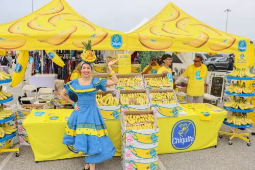 A woman in a blue dress and fruit headpiece poses in front of a Chiquita banana booth at a market. The booth, adorned with yellow decorations and multiple boxes of bananas, attracts several attendees, reminiscent of the bustling scene fueled by the $3.935 Million State Grant for Port of Hueneme's improvement.