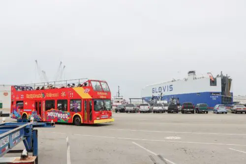 A red double-decker sightseeing bus with "Hollywood" branding is parked in front of a large car carrier ship labeled "GLOVIS" in an industrial port area, showcasing the bustling activity following the recent $3.935 million state grant for Port of Hueneme as Governor ramps up California Port Data Partnership Program.