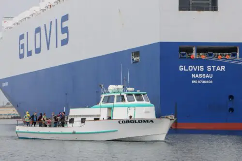 A small boat named Coroloma is sailing beside a large cargo ship labeled "GLOVIS STAR NASSAU" at a port. Multiple people are standing on the deck of the small boat, likely celebrating the recent $3.935 Million State Grant for Port of Hueneme as Governor Ramps Up California Port Data Partnership Program.