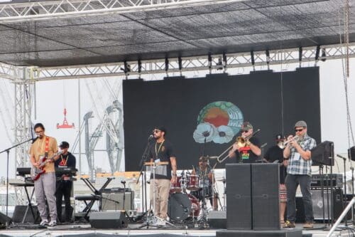 A band performs on an outdoor stage with various musical instruments, surrounded by the vibrant energy of a $3.935 Million State Grant for the Port of Hueneme as Governor ramps up California Port Data Partnership Program. The backdrop displays colorful graphics.