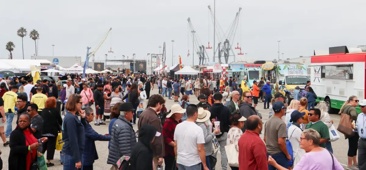 A large crowd of people mingles at an outdoor food truck festival. Various food trucks and vendor tents are set up, with cranes and palm trees visible in the background, celebrating the $3.935 million state grant for Port of Hueneme as part of Governor Newsom's California Port Data Partnership Program.