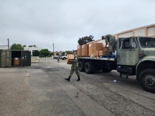 Military personnel in camouflage uniforms unload cardboard boxes from a truck in an open lot near storage containers, contributing to the $3.935 million State Grant for Port of Hueneme as Governor ramps up the California Port Data Partnership Program.