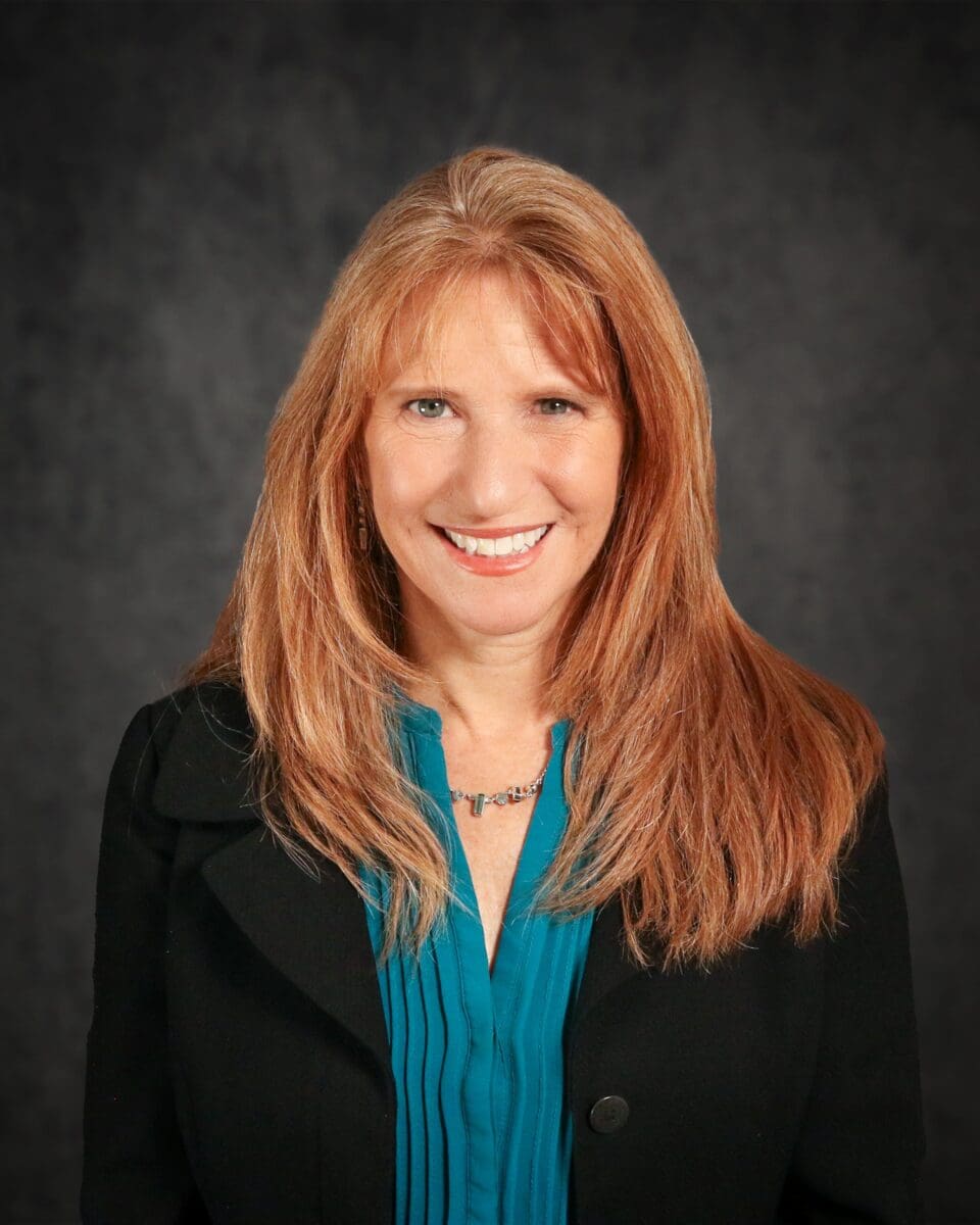 Stacy Lange, a woman with long red hair, smiles warmly in her turquoise blouse and black jacket against a dark gray background.