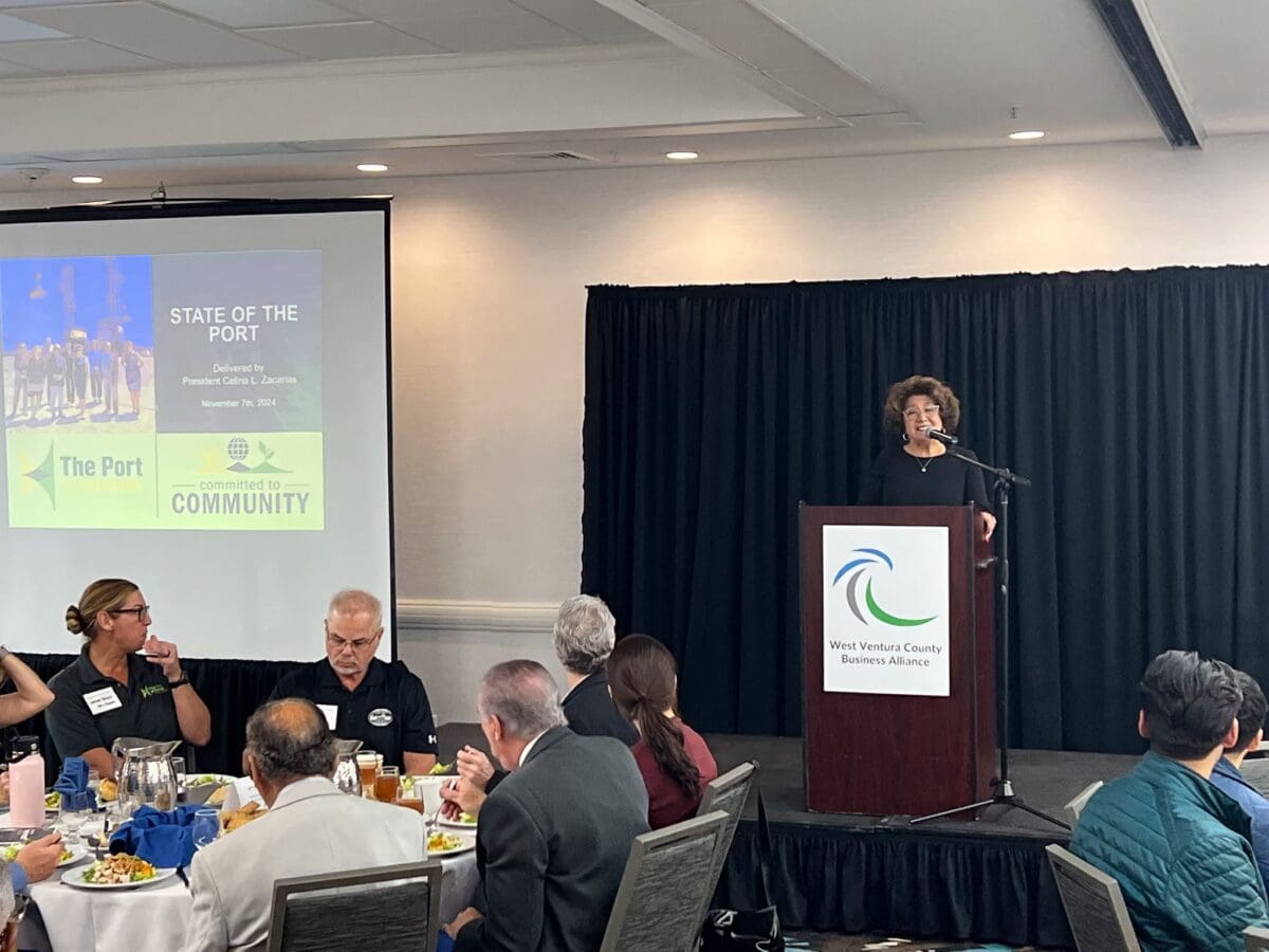 A speaker at a podium addresses attendees during a business event. Behind them, a projector displays "State of the Port" with news of the $3.935 Million state grant for Port of Hueneme. In the foreground, people sit at tables with food as California ramps up its Port Data Partnership Program.