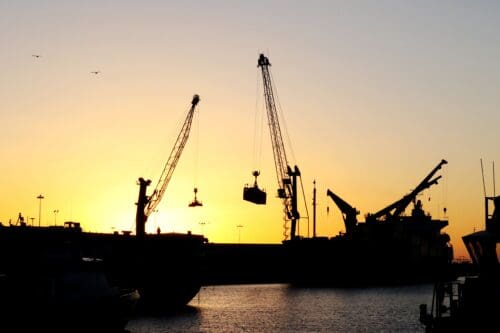 Silhouetted cranes and ships at a harbor during sunset, with a calm water surface and a few birds in the sky, set the scene as the Port of Hueneme embraces new horizons with a $3.935 million state grant under California's expanded Port Data Partnership Program.