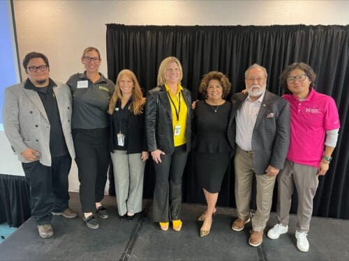 Seven people stand on stage, smiling at the camera in a mix of business and casual attire. Against a black curtain backdrop, they celebrate receiving the $3.935 million state grant for Port of Hueneme as part of the Governor's initiative to enhance California's Port Data Partnership Program.