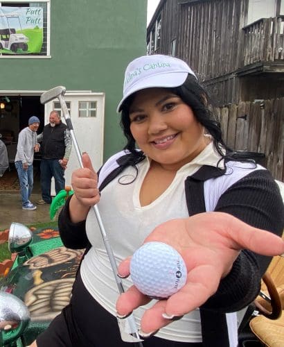 A person grins while holding a golf ball and putter on a bustling mini-golf course, reminiscent of the excitement surrounding the $3.935 million state grant for Port of Hueneme as California strengthens its port data partnership program.