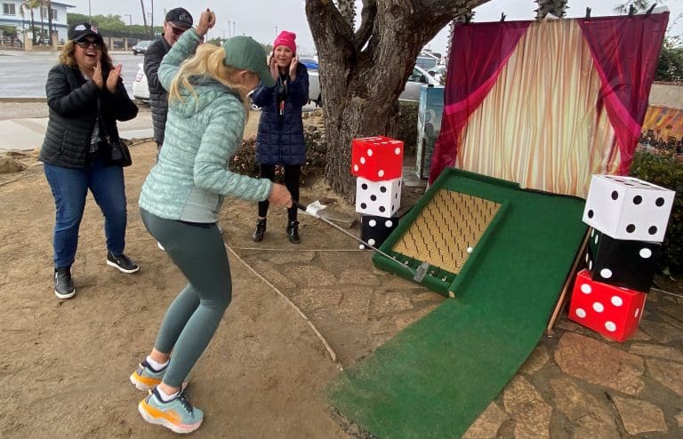 A woman plays mini-golf outdoors, watched by three people, as she puts towards a ramp adorned with large dice. Nearby, discussions buzz about the $3.935 million state grant for the Port of Hueneme, part of California's port data partnership program ramping up progress statewide.