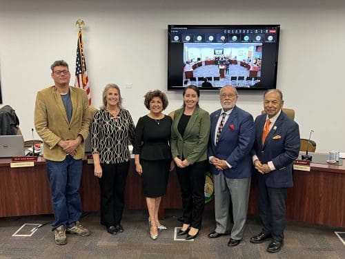 Six people stand in a meeting room with a large screen displaying a video call, the American flag to the left. Discussions likely swirl around the recent $3.935 million state grant for Port of Hueneme, as the Governor intensifies efforts on Californias Port Data Partnership Program.