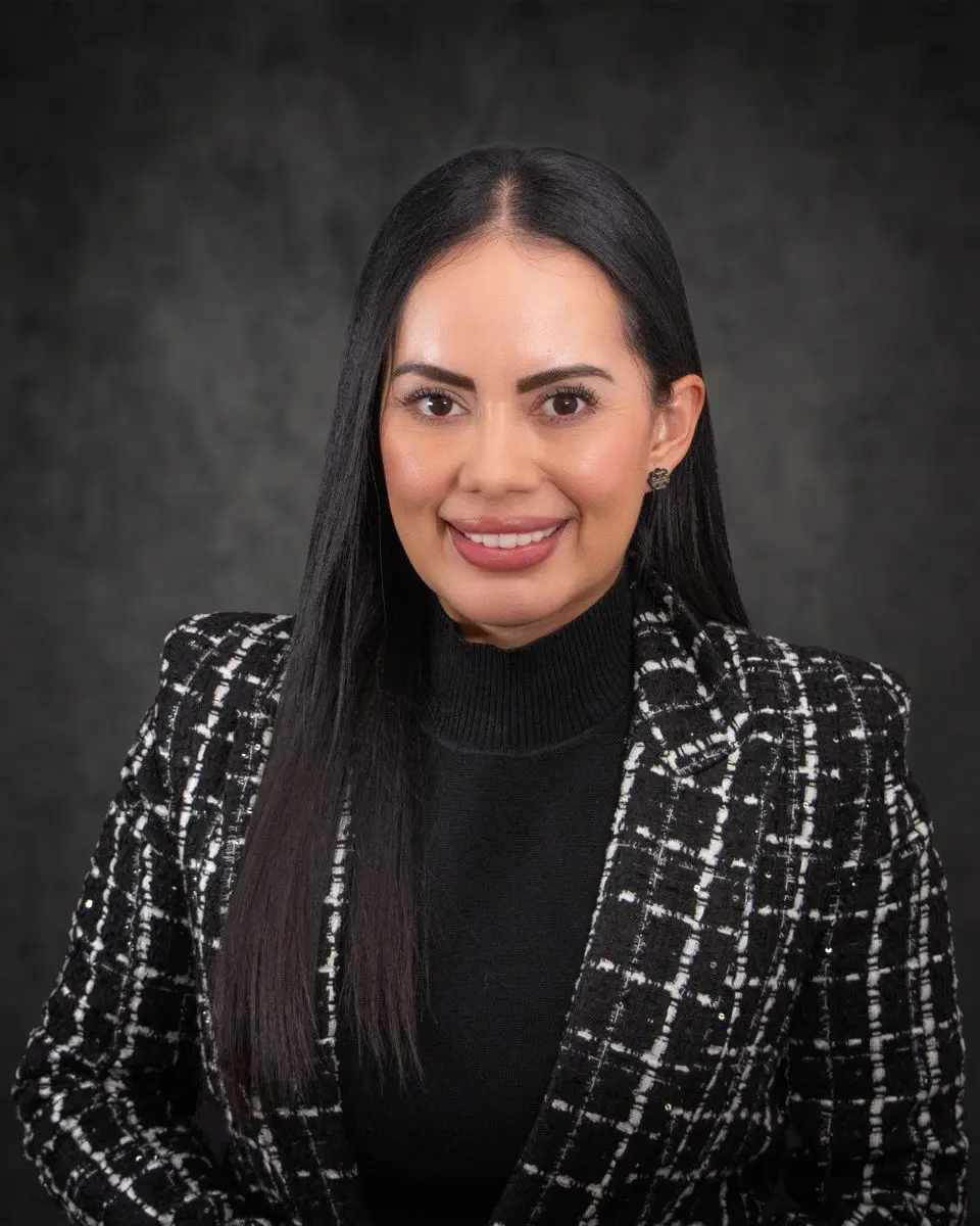 Mariana Hernandez, with her long dark hair, smiles warmly while wearing a black and white plaid jacket and a black turtleneck against a dark background.