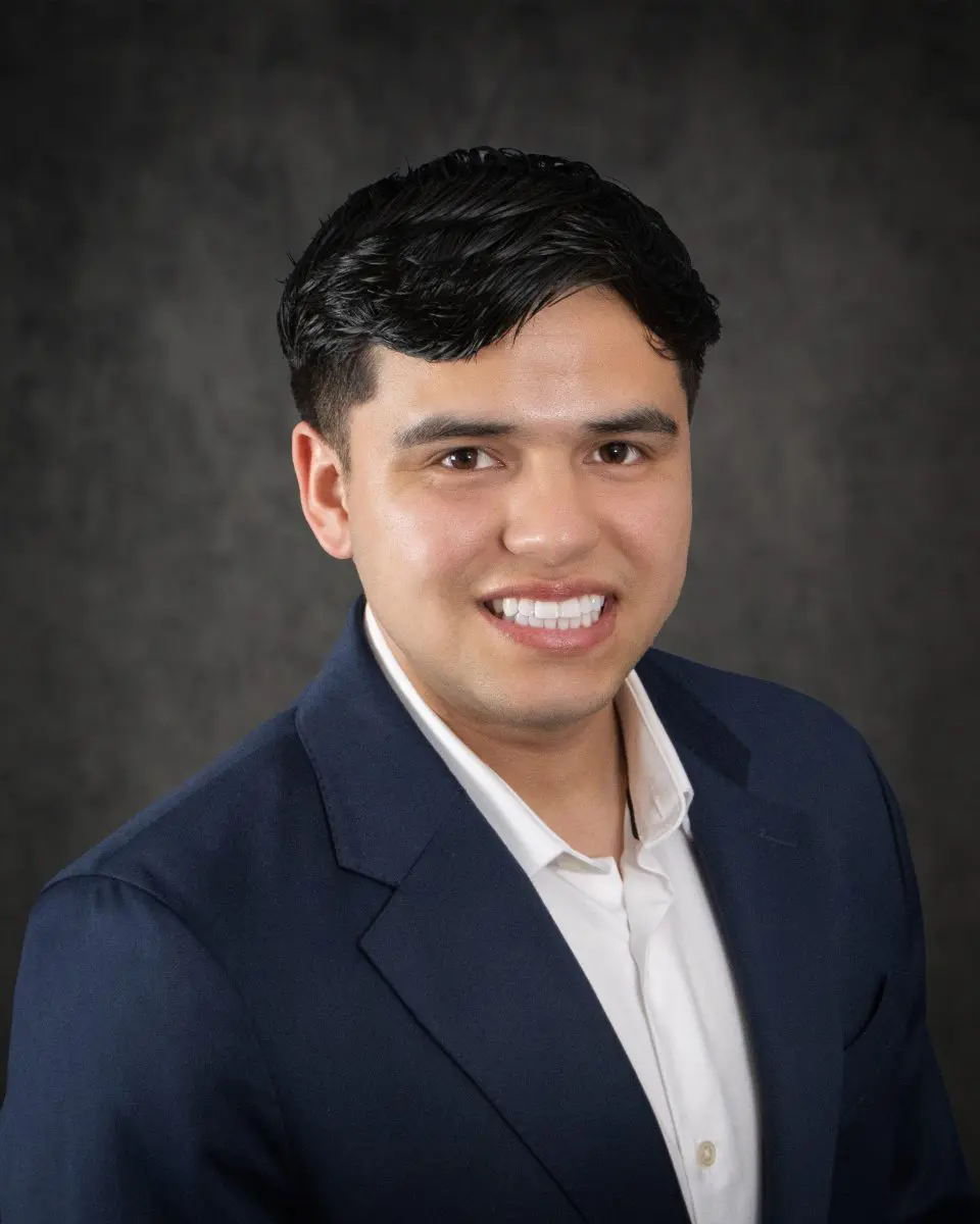 Ronaldo Esquivel, with his short dark hair, is wearing a white shirt and a dark suit jacket, smiling confidently against a plain dark background.