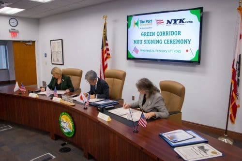 Three people sign documents at a conference table during the Green Corridor MOU Signing Ceremony. A screen displays details of the event, highlighting a $3.935 million state grant for the Port of Hueneme as the governor ramps up Californias port data partnership program. Flags and an emblem adorn the table.