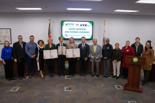 A group of people stands in a conference room, holding documents, as the screen behind them displays Green Corridor MOU Signing Ceremony. Flags and a podium are visible, celebrating the $3.935 Million State Grant for Port of Hueneme, as part of Californias port data partnership program.