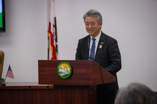 A person in a suit speaks at a podium adorned with a city seal, announcing the $3.935 Million State Grant for Port of Hueneme. The backdrop of flags symbolizes unity as the Governor ramps up Californias Port Data Partnership Program.