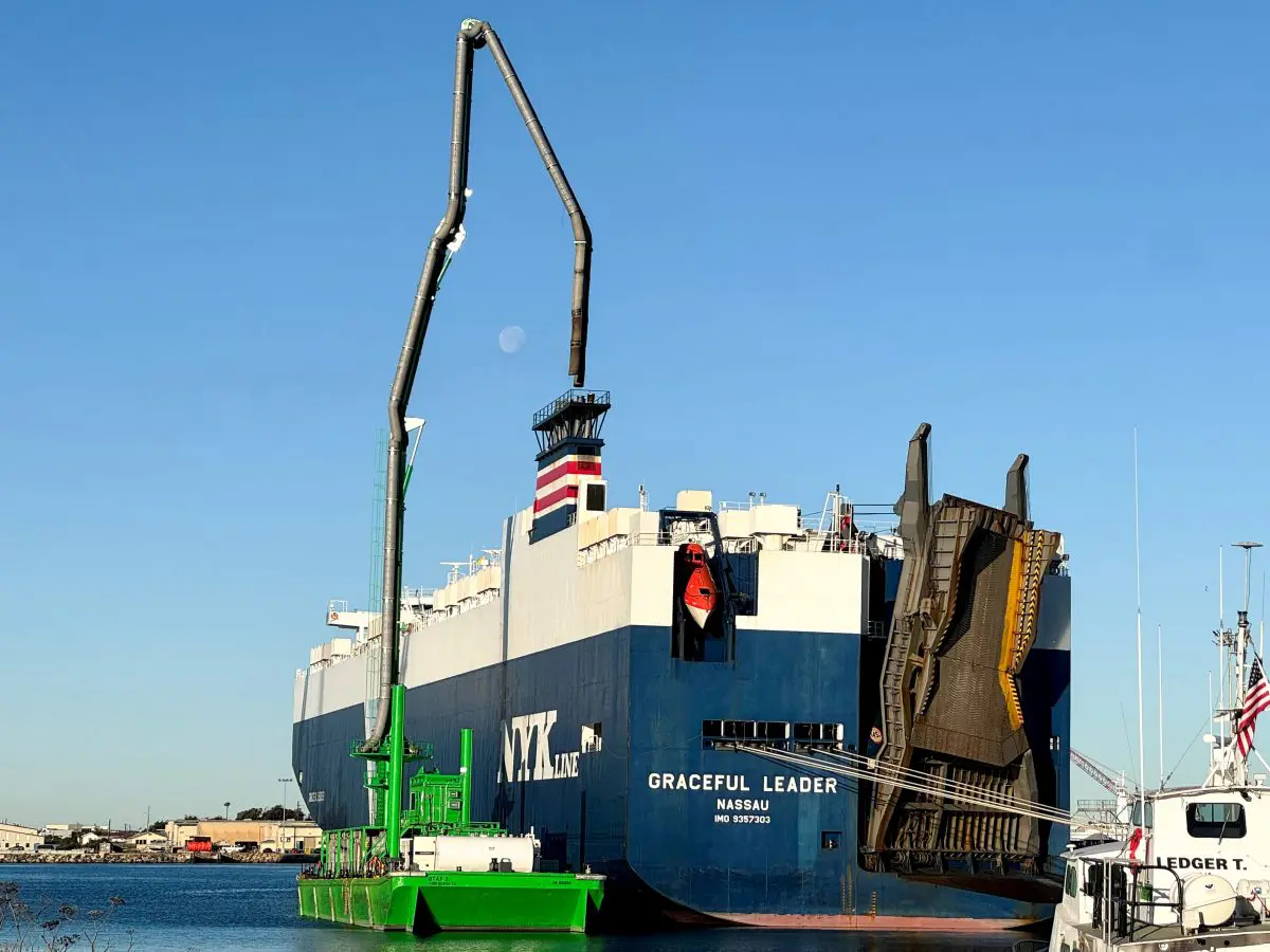 A large blue and white cargo ship is docked at the Port of Hueneme, sporting a crane on its deck. Under the clear blue sky, with the moon in view, this bustling port benefits from a $3.935 million state grant as part of Californias enhanced Port Data Partnership Program.