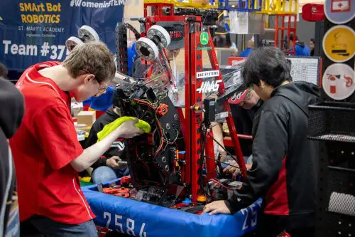 Three individuals work diligently on a robot at a competition, adjusting parts and wiring. Behind them, a banner with SMArt Bot Robotics stands tall, symbolizing innovation in a state where the $3.935 Million grant supports programs like California Port Data Partnership.