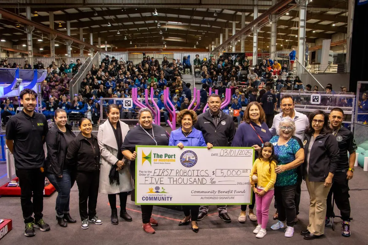 A group celebrates presenting a $5,000 check to FIRST Robotics, set against the backdrop of a cheering crowd and robotics event. The enthusiasm mirrors the excitement surrounding the $3.935 million state grant boosting California’s Port Data Partnership Program.