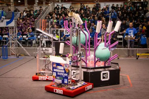 Robots from teams 2543 and 1388 compete in a thrilling robotics event, artfully maneuvering pink structures and teal balls as the large crowd watches in anticipation, all set against the backdrop of excitement around Port of Hueneme’s $3.935 million state grant announcement.