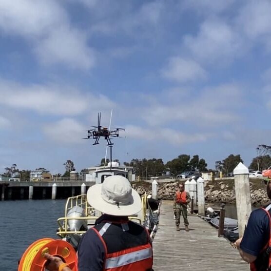 People in life vests standing on a coastal dock observe a drone hovering above water, with construction cranes and buildings in the background.