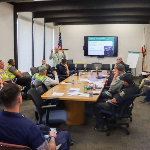 A group of people, some in uniforms and safety vests, are seated around a conference table in a meeting room with a presentation screen displaying information about the $3.935 Million State Grant for Port of Hueneme as Governor ramps up the California Port Data Partnership Program.