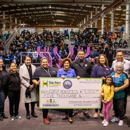 A group celebrates presenting a $5,000 check to FIRST Robotics, set against the backdrop of a cheering crowd and robotics event. The enthusiasm mirrors the excitement surrounding the $3.935 million state grant boosting California’s Port Data Partnership Program.