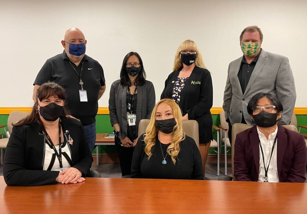 Seven people wearing face masks pose for a group photo in a room with a yellow and green accent wall, capturing the spirit of 2021. Four are standing in the back, and three are seated at a wooden table in front, as they gather for their year-end Dock Talk.