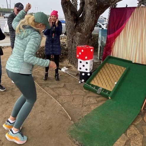 A woman plays mini-golf outdoors, watched by three people, as she puts towards a ramp adorned with large dice. Nearby, discussions buzz about the $3.935 million state grant for the Port of Hueneme, part of California's port data partnership program ramping up progress statewide.