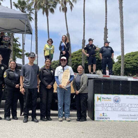 A group of people stands in front of a large check for $5,000, one person holding a wooden plaque. Police officers and bystanders are in the background against a backdrop of palm trees, celebrating the Port Hueneme Little League's generous donation.