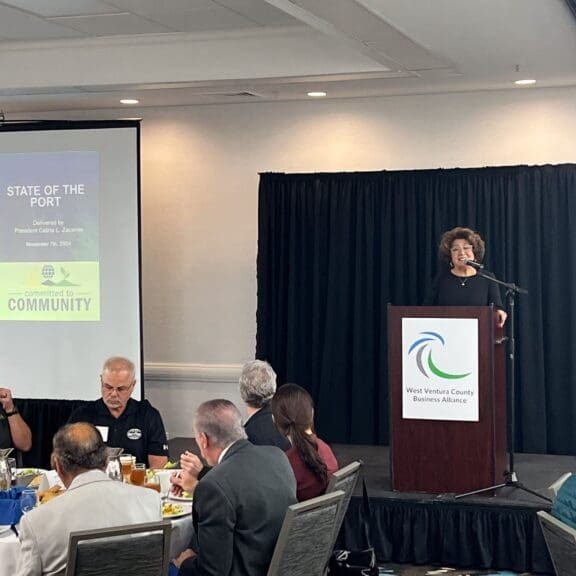 A speaker at a podium addresses attendees during a business event. Behind them, a projector displays "State of the Port" with news of the $3.935 Million state grant for Port of Hueneme. In the foreground, people sit at tables with food as California ramps up its Port Data Partnership Program.