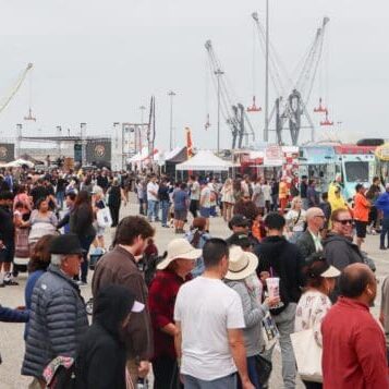 A large crowd of people mingles at an outdoor food truck festival. Various food trucks and vendor tents are set up, with cranes and palm trees visible in the background, celebrating the $3.935 million state grant for Port of Hueneme as part of Governor Newsom's California Port Data Partnership Program.