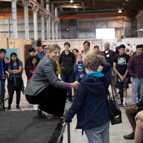 A woman kneels on a stage holding a microphone to a young boy in a blue jacket in front of a crowd, framed by the industrial-looking event space that feels oddly like home.